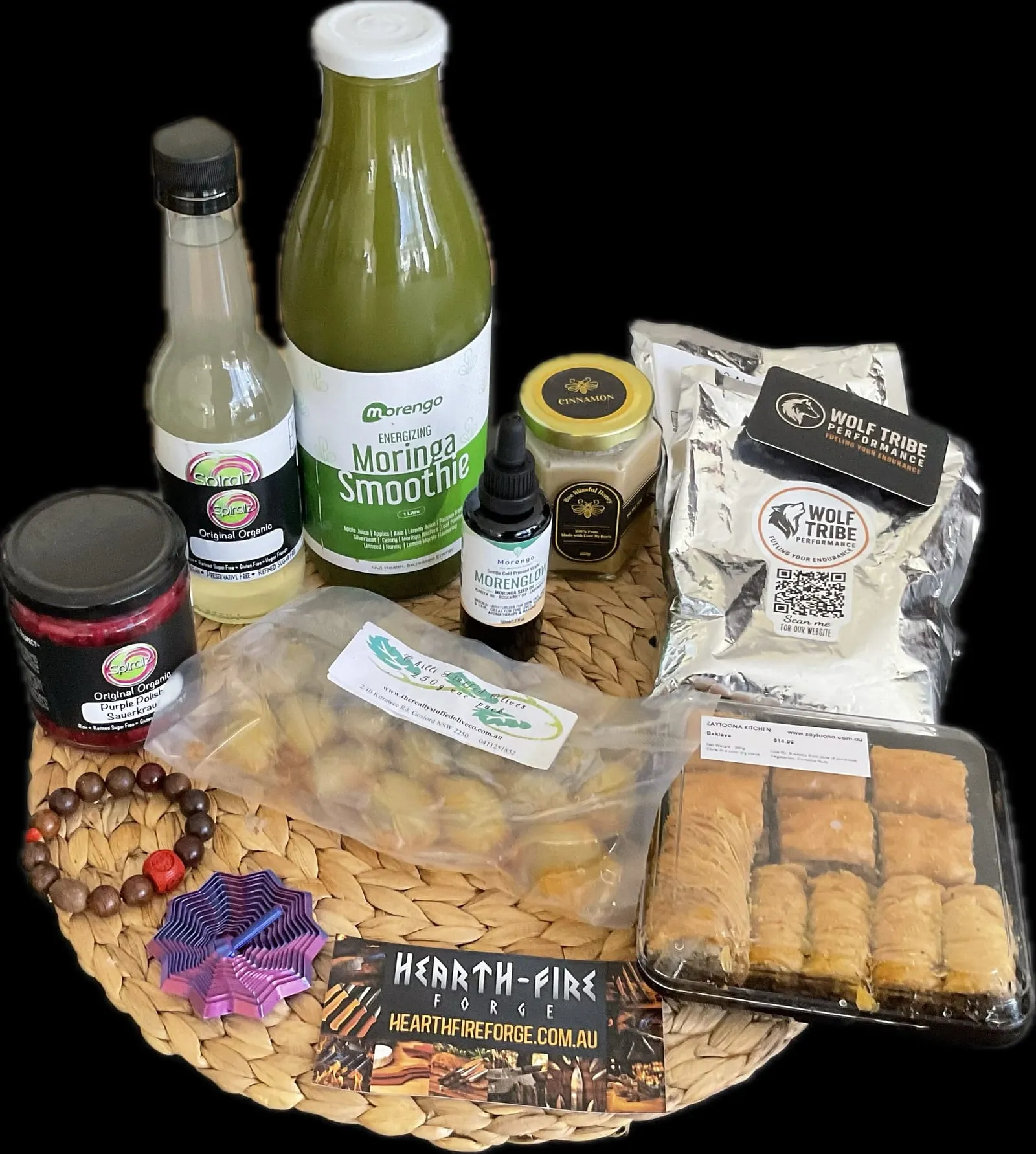 A haul of local goods from Gosford Markets laid out on a woven mat, including organic sauerkraut, a moringa smoothie, cinnamon honey, stuffed olives, baklava and a Hearth-Fire Forge business card.